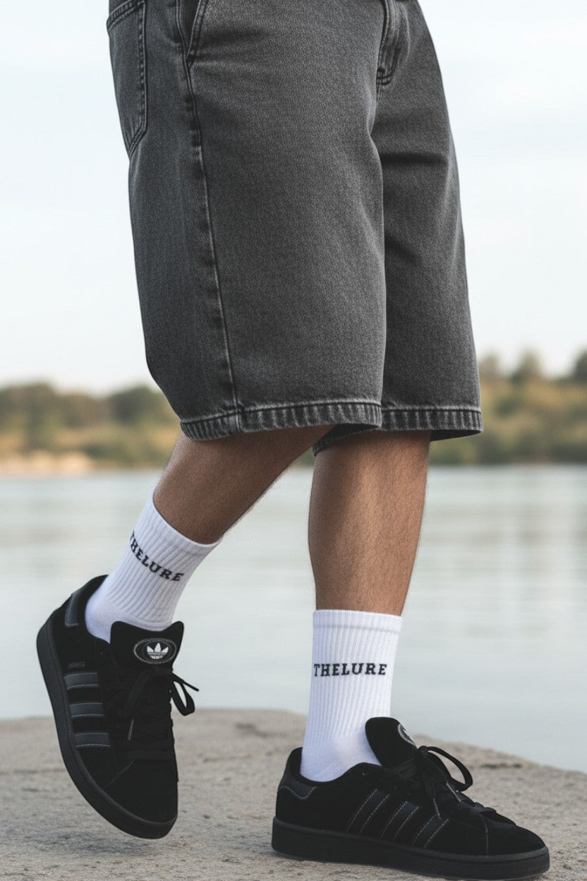 thelureclothing Performance Socke in Weiss mit Logo, getragen in schwarzen Sneakern und grauen Jeans-Shorts am Wasser.